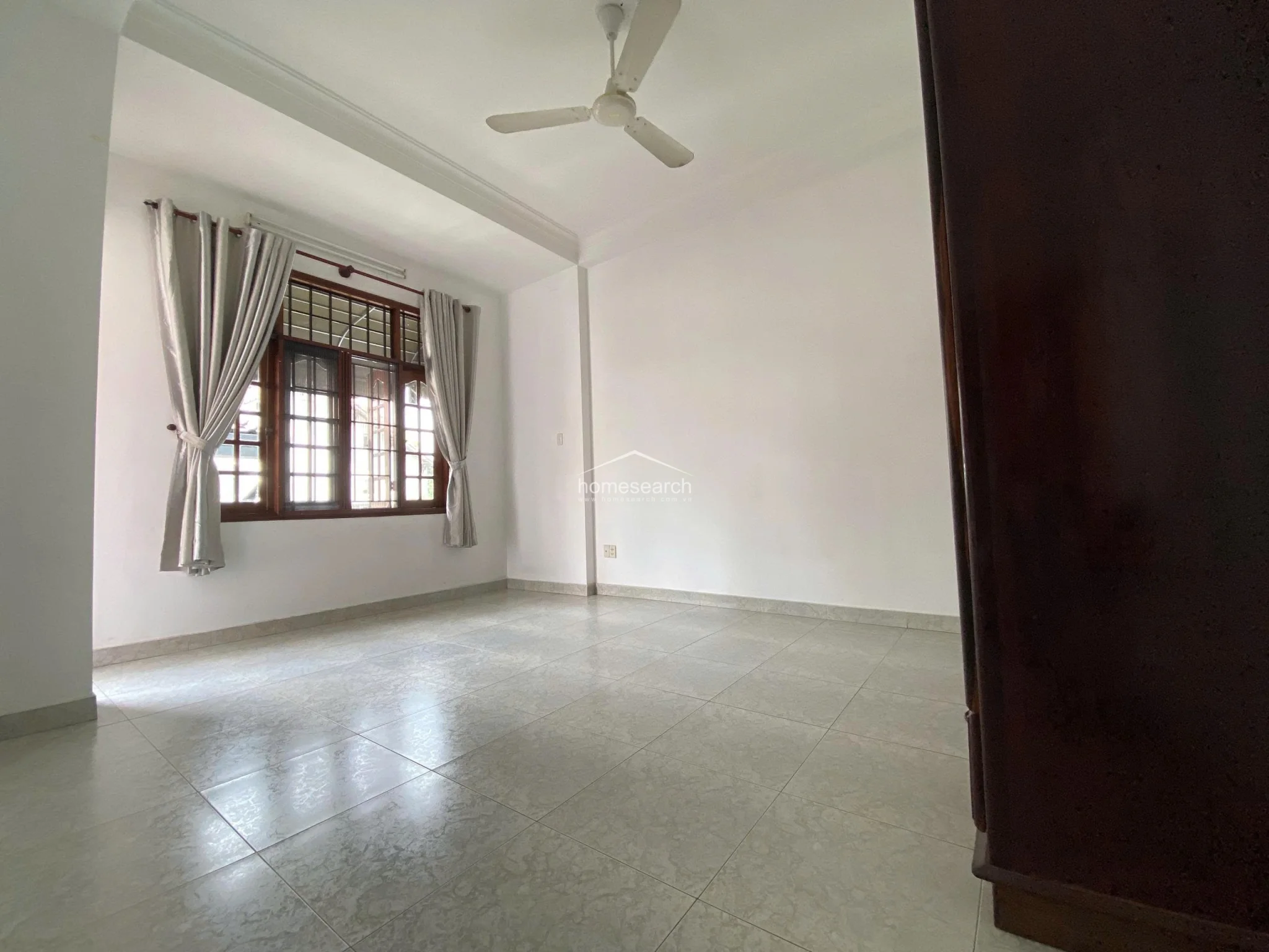 This bedroom or living space features light-colored floor tiles, white walls, and a ceiling fan for climate control. A large window with a dark wood frame and light curtains allows for significant natural light, while the room maintains a minimalist and well-kept aesthetic.