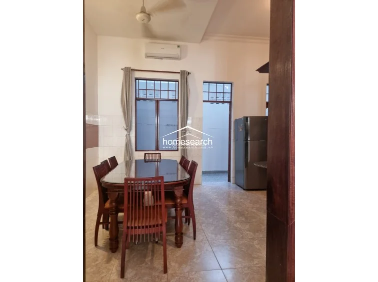 This dining room features a formal wooden table set and is illuminated by natural light from a large window dressed with simple curtains. The space is climate-controlled with a wall-mounted AC unit and includes a clear view into an adjacent kitchen area, which houses a large refrigerator.