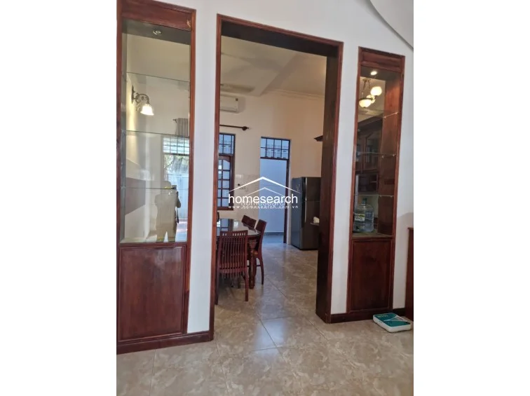 This image shows a transition area featuring custom-built, floor-to-ceiling wooden display cabinets with glass shelving flanking a wide doorway. The space features neutral, light-colored tile flooring and white walls, creating an open flow that leads from the foreground into a dining area and kitchen beyond.