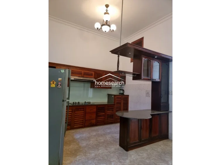 The kitchen features a traditional design with warm-toned wooden cabinetry, louvered cabinet doors, and a stone countertop. It is equipped with a built-in gas stovetop, a range hood, and a unique, kidney-shaped breakfast bar, all illuminated by a decorative chandelier under high ceilings.