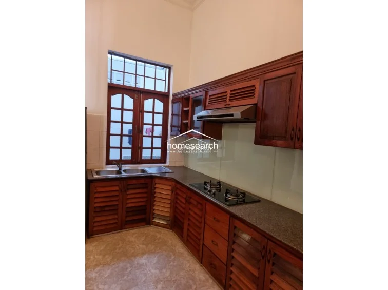 This corner kitchen layout maximizes utility with a large, multi-paned wooden window that provides natural light and ventilation. The space is well-maintained, featuring matching louvered wooden cabinetry, a stainless steel double sink, and a sleek glass-top gas stove set against a light-colored backsplash.