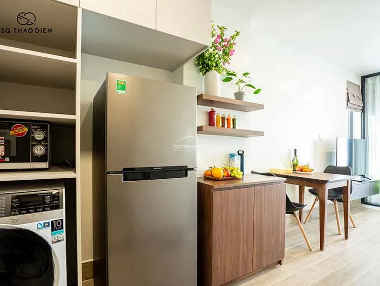 This functional kitchen and storage corner maximizes space with a tall, integrated shelving unit that houses a microwave and a washing machine. The inclusion of a stainless steel refrigerator and open wooden shelving provides a clean, organized look that complements the overall modern interior design.
