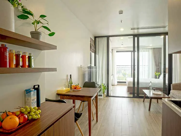 This view highlights the seamless flow between the dining area and the bedroom, emphasizing the airy, open feel created by the glass walls. The dining set is positioned conveniently near the kitchen, making it a practical space for meals, while the warm wood tones and soft lighting create a welcoming atmosphere.