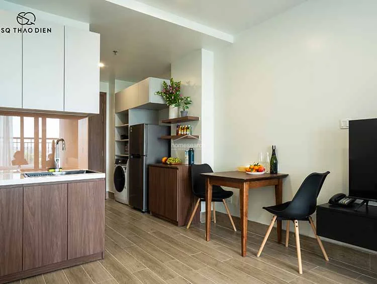 The kitchen is efficiently designed with a compact linear layout, featuring wood-grain cabinetry, white upper storage, and a light-colored countertop. It is well-equipped with a stainless steel refrigerator, a built-in washing machine, and floating shelves that provide stylish, accessible storage for kitchen essentials.