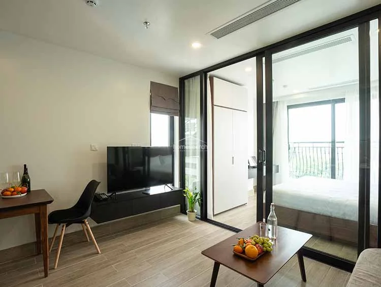 This living area features a modern, open-plan layout with wood-look tile flooring and neutral white walls that enhance the sense of space. A large sliding glass partition separates the living room from the bedroom, allowing natural light to flow through while maintaining a sleek, contemporary aesthetic.