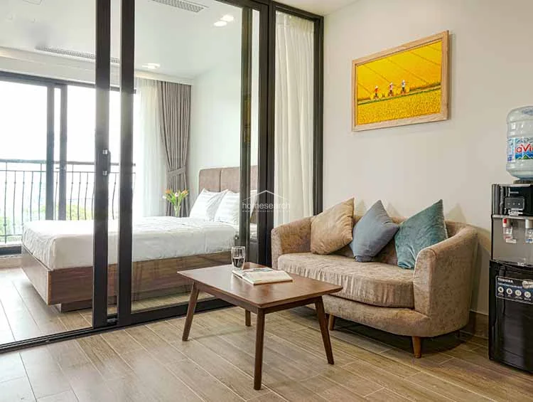 The living room includes a comfortable sofa, a wooden coffee table, and a wall-mounted television, creating a cozy and functional relaxation zone. A large window with a roman shade provides natural light, and the space is accented with a vibrant piece of wall art and a convenient water dispenser.