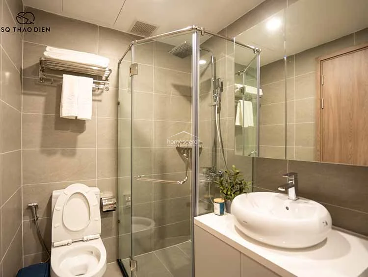 This compact, modern bathroom is finished in neutral grey tiles and features a glass-enclosed corner shower stall for efficient space utilization. The vanity includes a white vessel sink atop a sleek cabinet, complemented by a large mirror that enhances the sense of space.