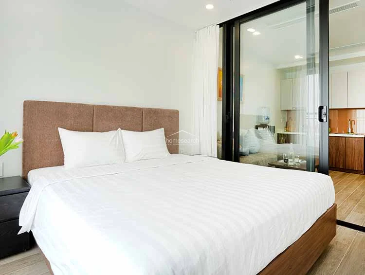 This bedroom features a clean, modern aesthetic with a plush brown upholstered headboard and light wood-tone flooring. It connects directly to the kitchen area through a sleek, black-framed glass sliding door, allowing for an open-concept flow and shared natural light.