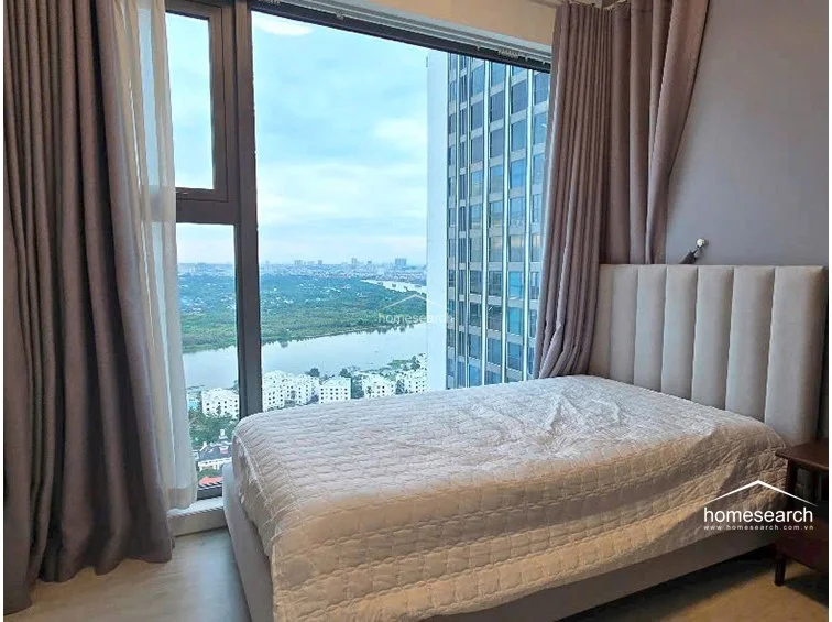 This bedroom features a floor-to-ceiling glass window that provides an expansive, panoramic view of the river and surrounding cityscape. The room is furnished with a modern, upholstered bed frame and is finished with light-toned wood flooring and soft grey wall accents.