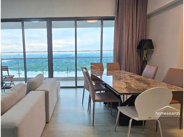 This open-concept living and dining area features floor-to-ceiling glass sliding doors that offer expansive, unobstructed river and city views. The space is furnished with a contemporary marble-top dining table and a modular light-grey sofa, complemented by neutral wall tones and light-colored wood flooring.