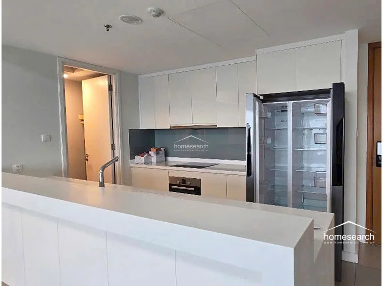 The kitchen layout includes a central island or breakfast bar that connects the cooking zone to the rest of the living space, promoting an open and social flow. It is well-equipped with a large, modern stainless steel refrigerator and ample storage within the handle-less white cabinets, reflecting a high standard of maintenance and contemporary design.