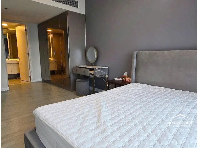 The bedroom layout includes a dedicated vanity area with a round mirror and a built-in, floor-to-ceiling wardrobe with a sleek, reflective finish. The space flows directly into an en-suite bathroom, which is visible through the open doorway, maintaining a cohesive and contemporary design aesthetic.
