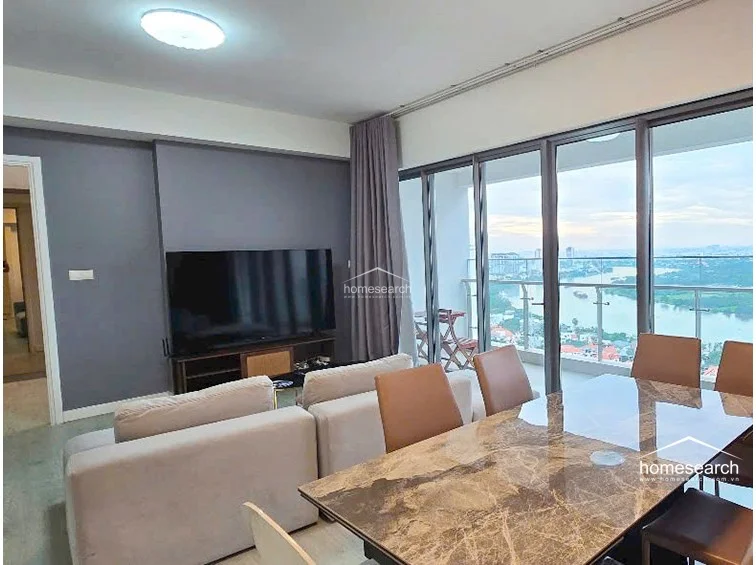 The dining area is anchored by a sophisticated marble-topped table and modern chairs, positioned to maximize the panoramic water views through the glass balcony doors. The neutral color palette and clean lines contribute to a high-end, contemporary feel, with heavy floor-length curtains providing privacy when needed.