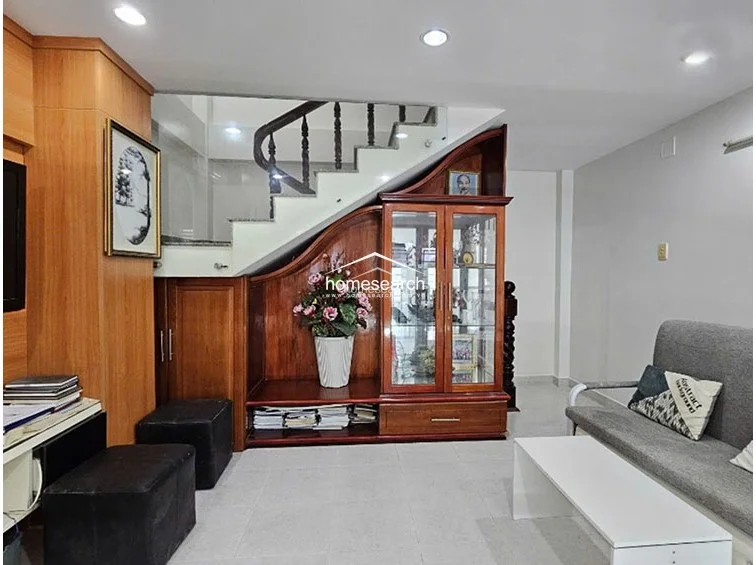 This living area is designed with a neutral color scheme and features custom-built wooden cabinetry integrated neatly under the staircase. The space is furnished with a grey sofa and a low-profile white coffee table, maintaining a clean and organized aesthetic.