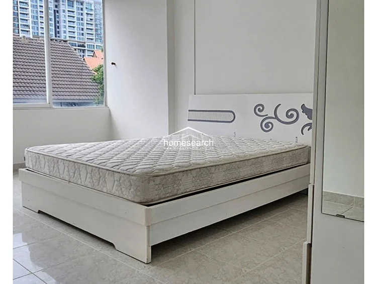 This bedroom is minimalist and bright, featuring a white bed frame and a large window that offers an urban view of neighboring buildings. The room is finished with light-colored floor tiles and plain white walls, providing a neutral canvas that maximizes the natural light.