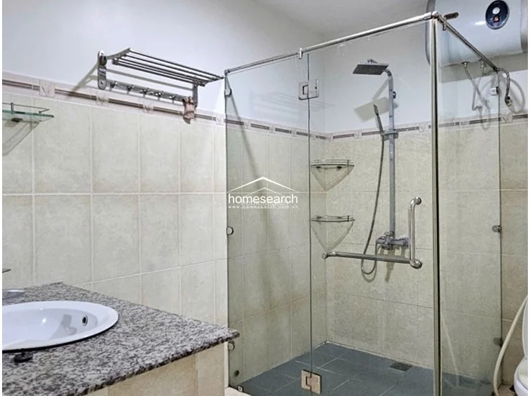 The bathroom is equipped with a modern glass-enclosed shower stall, a granite-topped vanity with a white basin, and a wall-mounted water heater. The walls are finished in neutral beige tiles with a decorative border, and the overall condition appears well-maintained and functional.