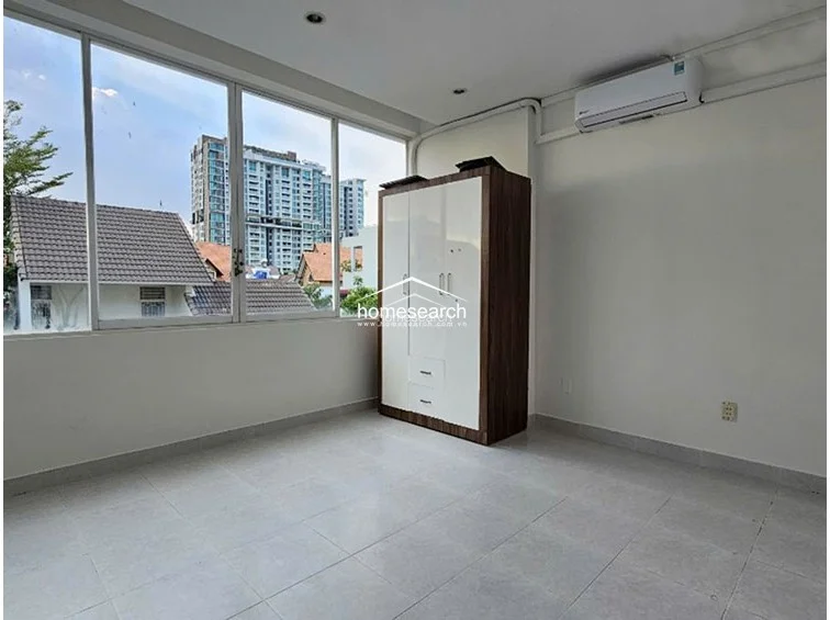 This bedroom is defined by its expansive floor-to-ceiling windows that provide abundant natural light and an urban view of the surrounding neighborhood. It includes a freestanding two-tone wardrobe for storage, a wall-mounted air conditioning unit, and simple, neutral finishes throughout.