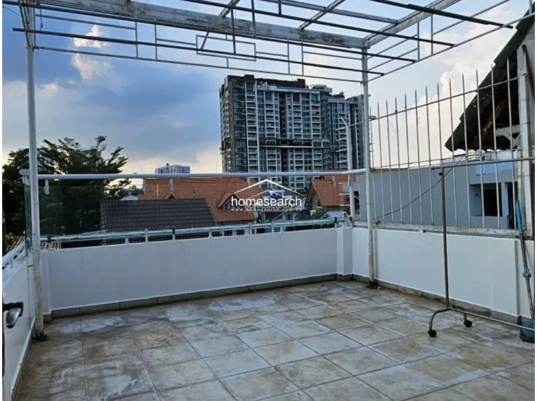 This rooftop terrace offers an open-air space with tiled flooring and a metal pergola frame, perfect for outdoor relaxation or laundry drying. The area is secured with glass-paneled railings and metal fencing, providing clear views of the adjacent high-rise buildings and local rooftops.