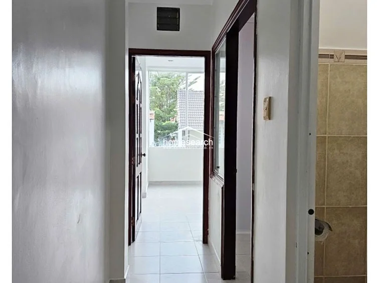 This interior hallway features neutral white walls and light-colored floor tiles, creating a clean and bright transition between rooms. The space connects to a bathroom on the right and leads directly into a sunlit room with large windows at the far end.