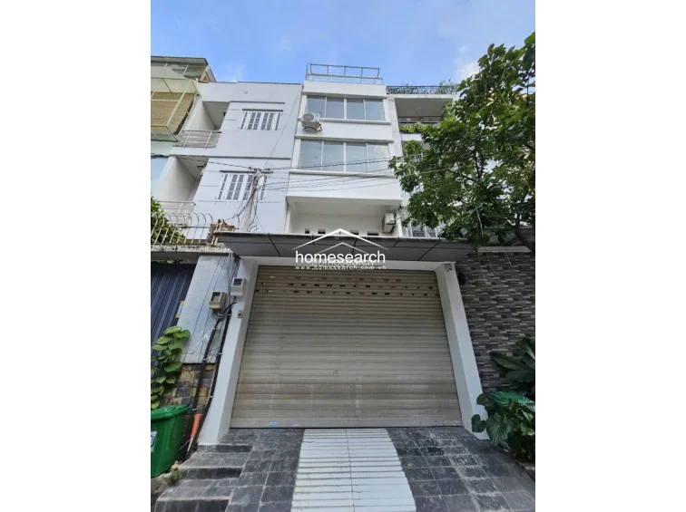 This is the exterior facade of a multi-story townhouse featuring a roll-up metal garage door at ground level and multiple levels of windows above. The building has a modern, functional design with an external AC unit visible, and it is situated on a paved street with some greenery nearby.