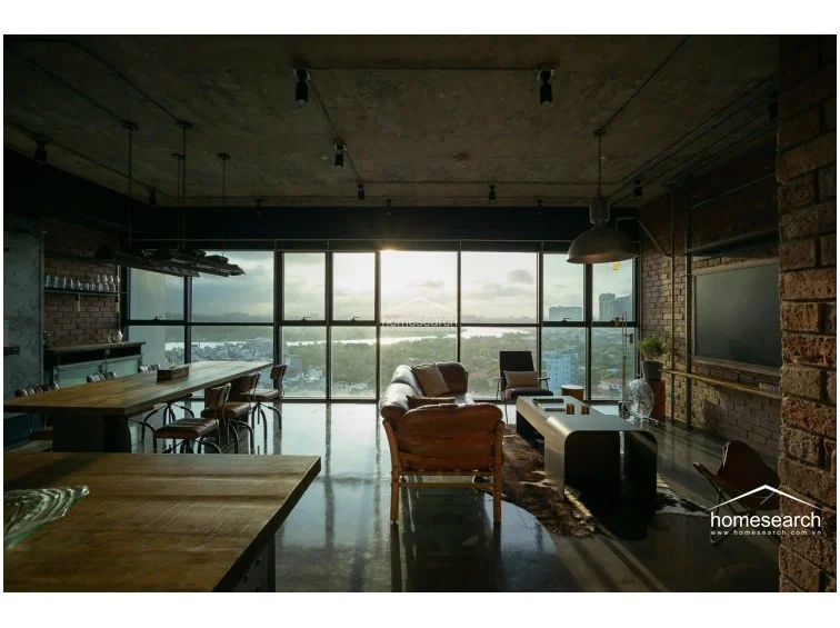 This expansive open-concept living and dining area features a striking industrial aesthetic with exposed concrete ceilings, brick walls, and polished concrete flooring. Floor-to-ceiling windows span the entire wall, offering panoramic city and river views while flooding the space with natural light. The room is furnished with premium leather seating and a large rustic wooden dining table, complemented by custom industrial-style pendant lighting.