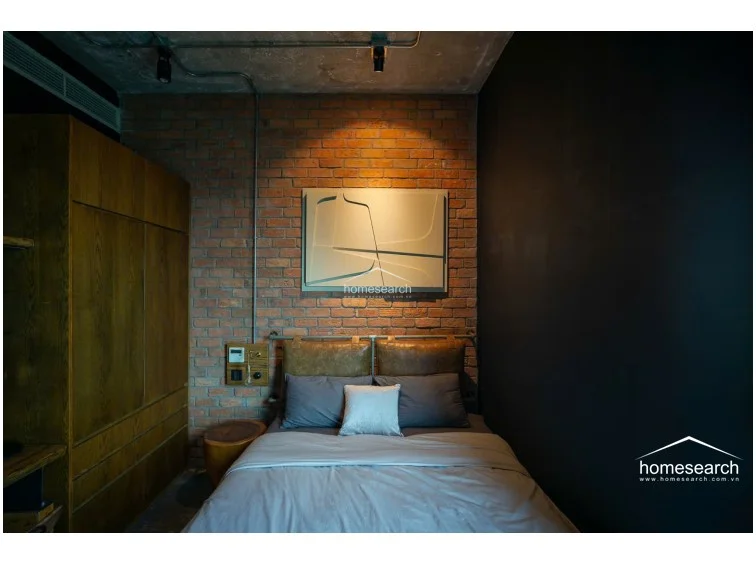 This industrial-style bedroom features a striking black color scheme with exposed concrete ceilings and metal conduit detailing. A unique glass-paneled wall provides a visual connection to the bathroom, while a metal wardrobe and integrated desk area maximize storage and functionality in a compact footprint.
