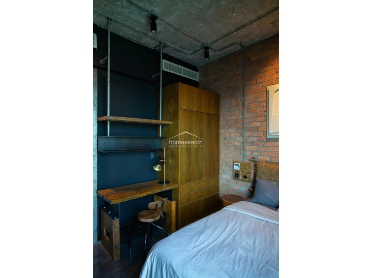 This bedroom offers an expansive, unobstructed view of the cityscape through floor-to-ceiling corner windows, flooding the space with natural light. The room is anchored by a plush, tufted headboard and a decorative wooden wall art piece, creating a sophisticated contrast against the dark, moody interior palette.