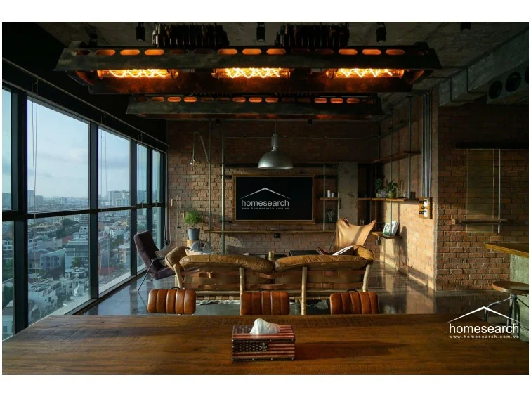 The living area is anchored by a comfortable brown leather sofa and a unique metal coffee table, perfectly positioned to enjoy the expansive exterior views. Exposed brick walls and visible conduit piping emphasize the loft-style architecture, while open shelving provides a curated space for decor. The overall maintenance appears excellent, with a cohesive design that balances raw industrial textures with warm, inviting furniture.