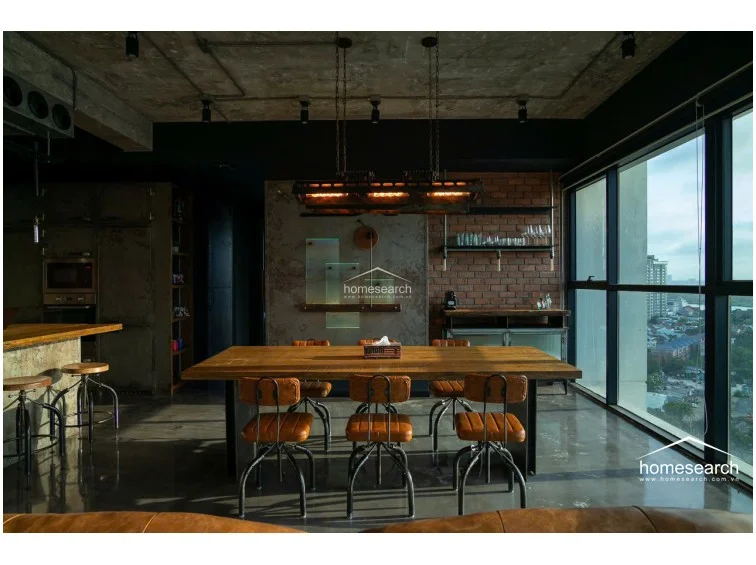 This view highlights the seamless flow between the dining area and the kitchen, characterized by a large wooden table and matching leather-upholstered stools. The kitchen features a robust island with a riveted metal finish and integrated appliances, including a wall-mounted oven and microwave. The space is well-lit by dramatic, custom-designed industrial light fixtures that hang over the dining table, creating a sophisticated focal point.