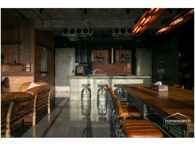 The kitchen layout centers around a functional island, which serves as both a workspace and a breakfast bar with industrial-style adjustable stools. The cabinetry and appliances are integrated into a sleek, metallic-finished unit, contrasting beautifully against the warm, textured brick backsplash. The open floor plan ensures an unobstructed view across the living area, maintaining a spacious and airy atmosphere.