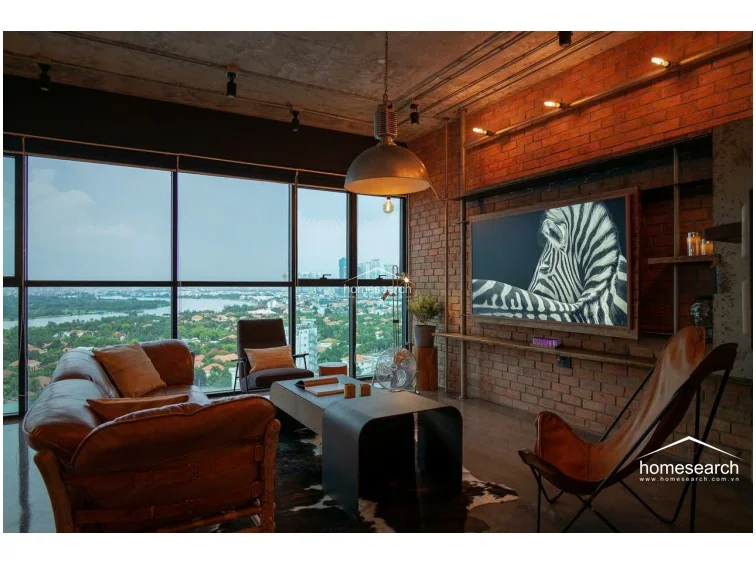 This industrial-style living room features floor-to-ceiling windows that provide expansive panoramic views and abundant natural light. The space is anchored by a rich leather sofa and a unique butterfly chair, set against a backdrop of exposed brick walls and polished concrete flooring.