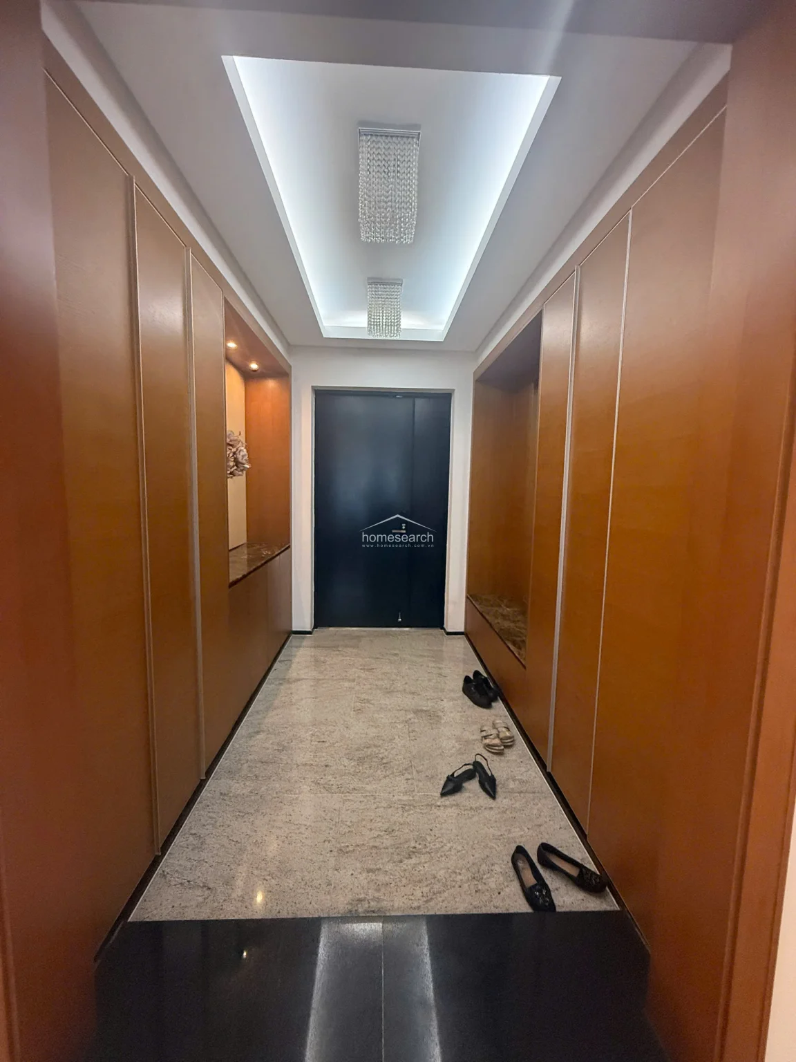 This entryway features extensive, floor-to-ceiling wooden cabinetry that provides ample concealed storage, complemented by a dedicated recessed bench area. The space is finished with polished stone flooring and sophisticated dual crystal chandeliers, creating a luxurious and welcoming first impression.