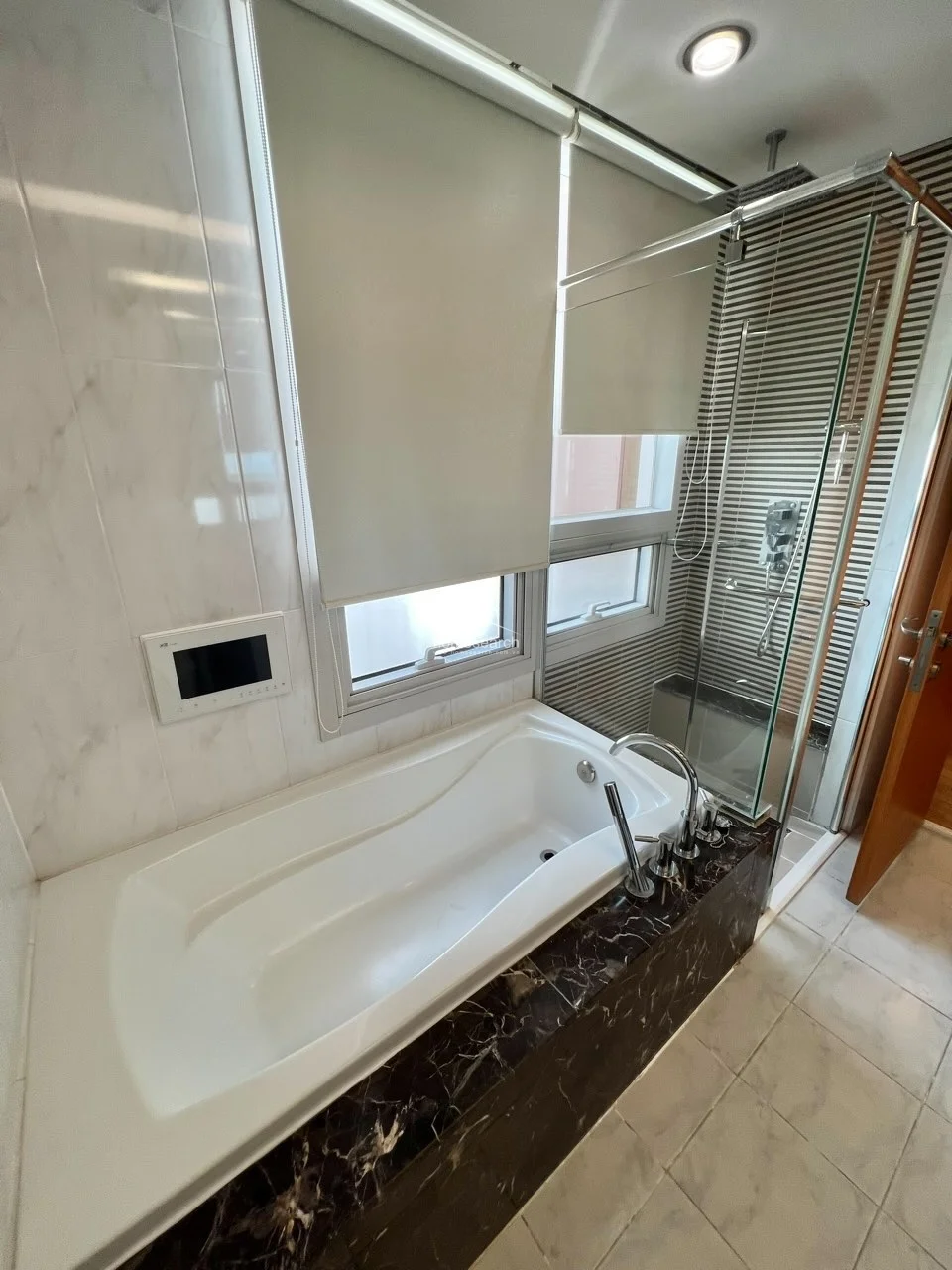 This bathroom is designed with a luxurious aesthetic, featuring a deep soaking tub encased in dark, high-contrast marble-look tile. A separate glass-enclosed shower stall with horizontal-striped wall accents adds a modern architectural touch, while the large window with roller shades ensures privacy and natural light. The space is well-appointed with chrome fixtures and includes a wall-mounted digital control panel, reflecting a high level of maintenance and contemporary design.