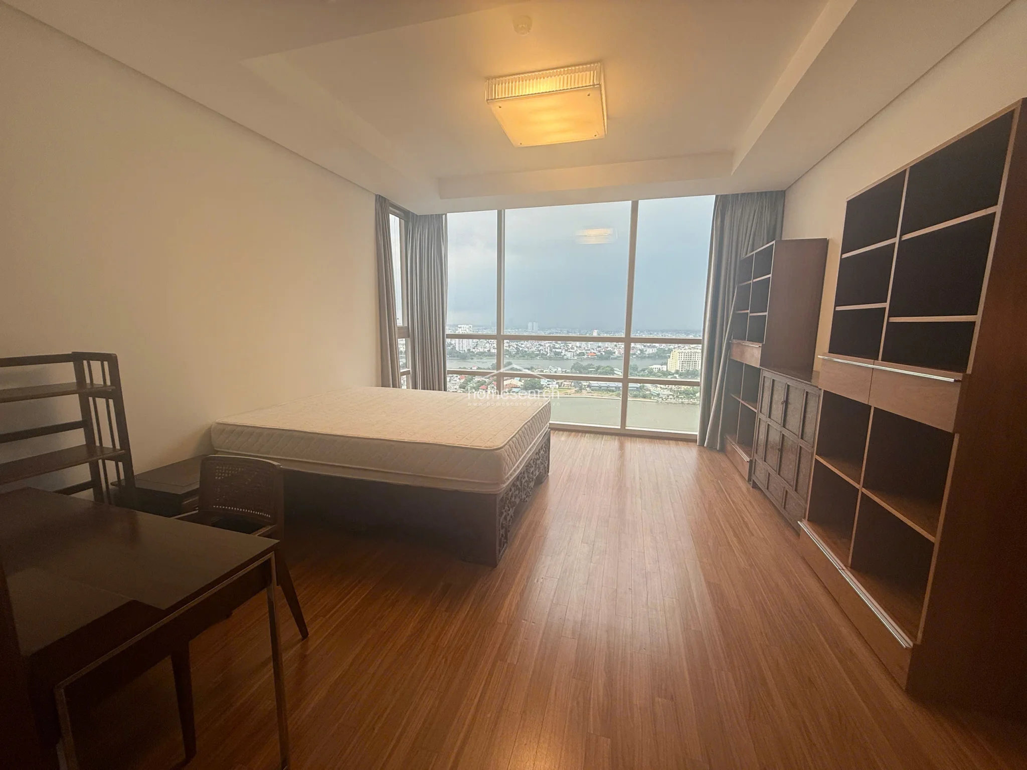 This view highlights the bedroom's generous storage capacity, featuring a tall, open-concept wooden shelving unit that complements the room's warm aesthetic. The layout remains open and airy, centered around the bed which faces the impressive floor-to-ceiling window wall. The overall condition appears well-maintained, emphasizing a functional and uncluttered living environment.