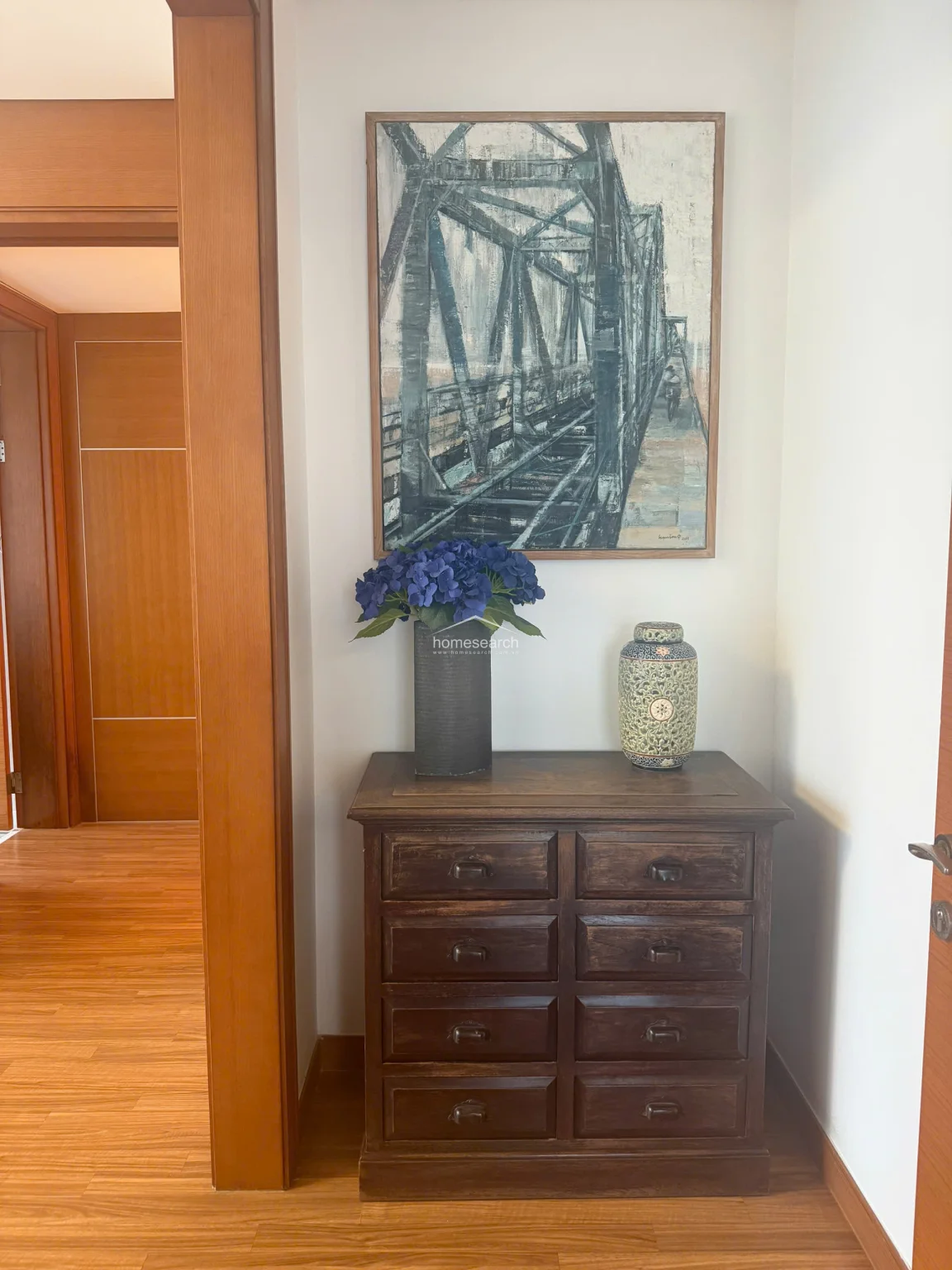 This elegant entryway or hallway vignette features a classic dark wood chest of drawers topped with a decorative vase and a vibrant blue floral arrangement. A large, framed painting of an industrial bridge hangs above, adding a sophisticated architectural focal point that transitions into the adjacent wood-floored living area.