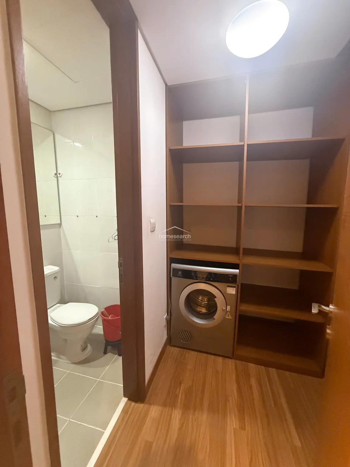 This utility area serves as a transition space, housing a built-in Electrolux front-loading washing machine integrated into a tall, multi-shelf wooden storage unit. The space connects directly to a tiled bathroom, which is finished in white wall tiles and features standard plumbing fixtures. The flooring transitions from the wood-look laminate of the main living area to practical, easy-to-clean ceramic tile in the wet zone.