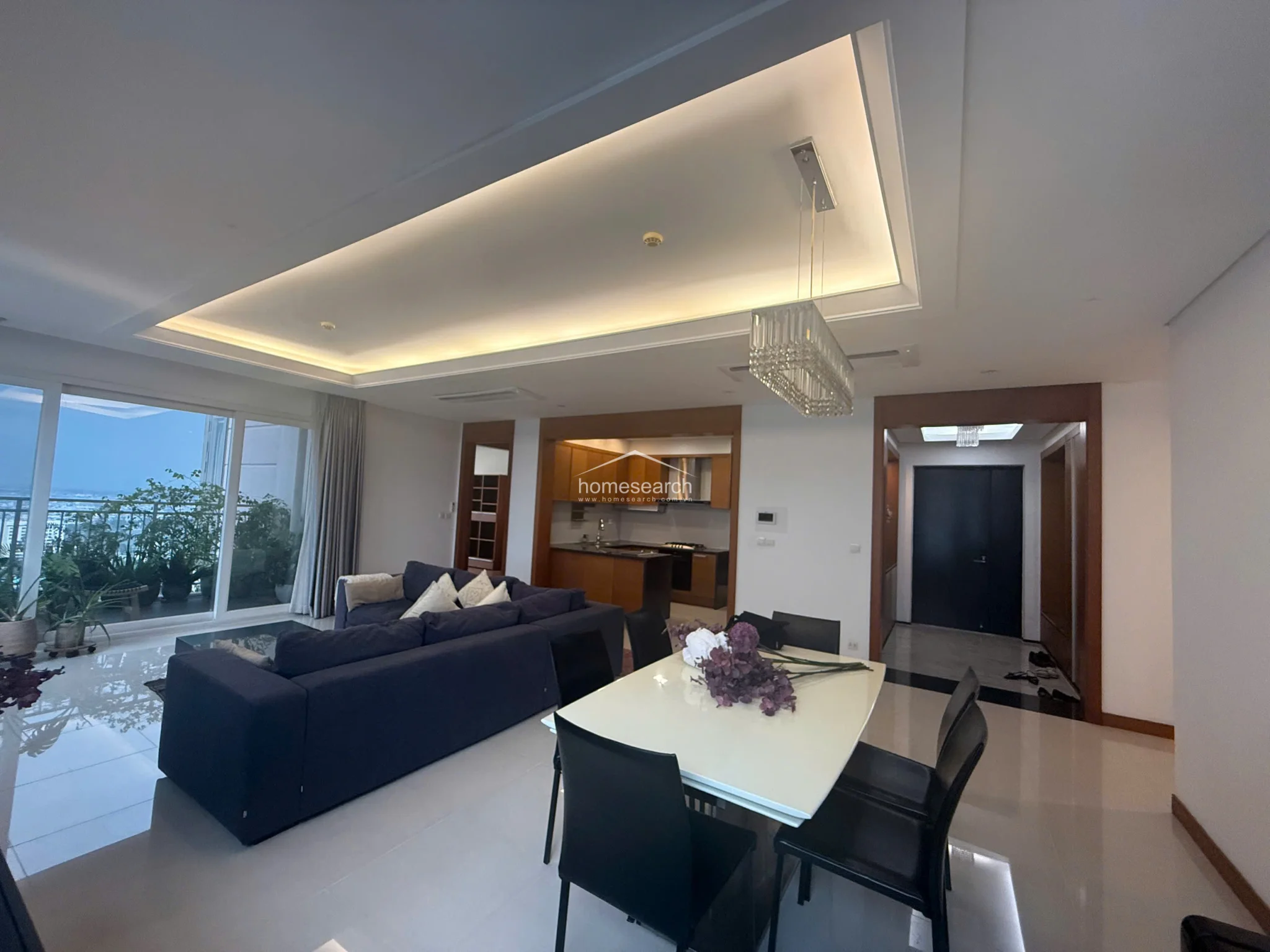 This open-concept living and dining area features polished light-colored tile flooring and a sophisticated tray ceiling with recessed ambient lighting. The space flows seamlessly into a kitchen with warm wood cabinetry and connects to an outdoor balcony through large glass sliding doors that provide ample natural light and city views.