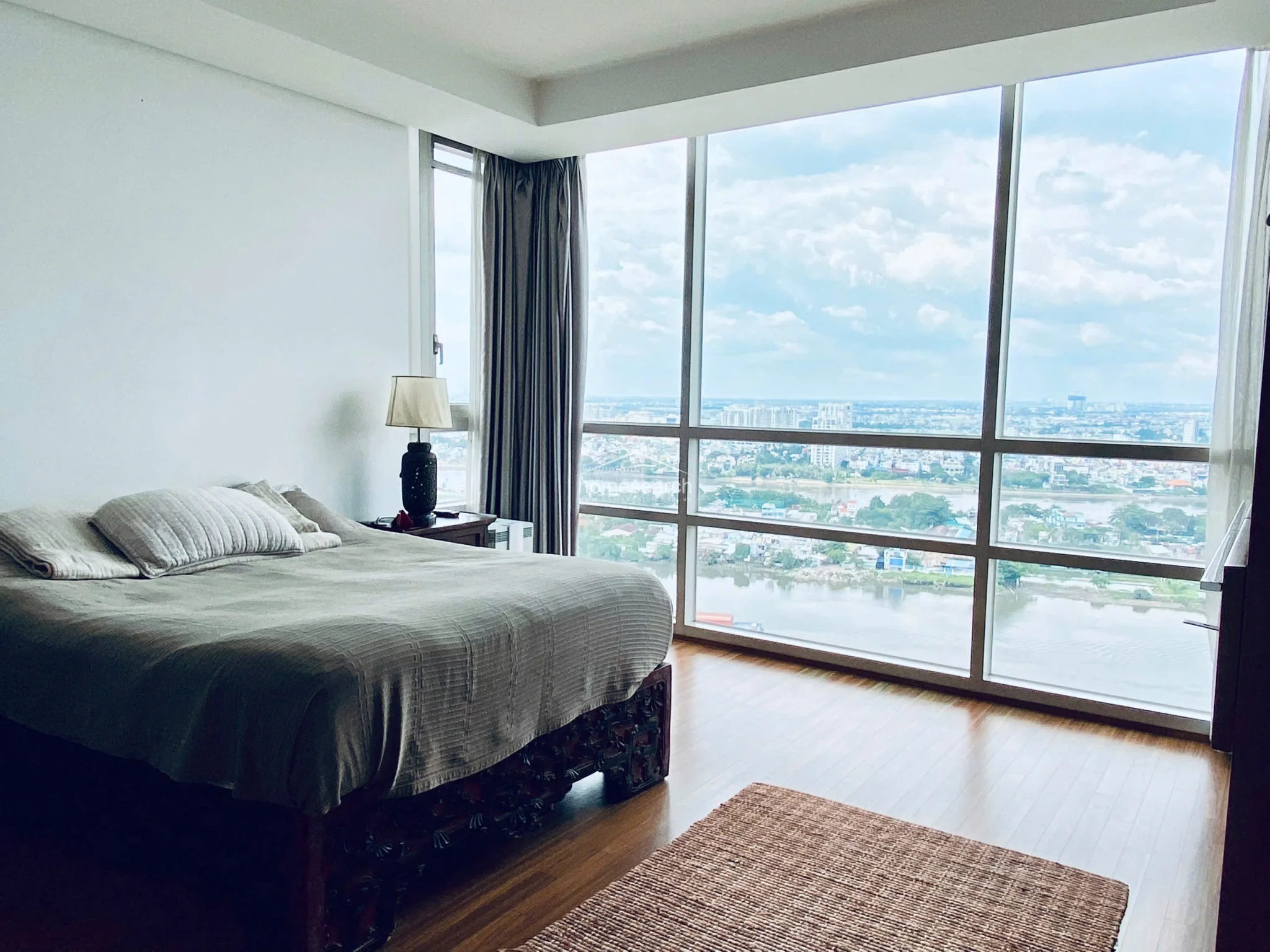 This primary bedroom features stunning floor-to-ceiling corner windows that provide panoramic river and city views, flooding the space with natural light. The room is finished with warm wood-tone flooring, white walls, and a distinctive carved wooden bed frame, complemented by a simple bedside lamp and neutral bedding.