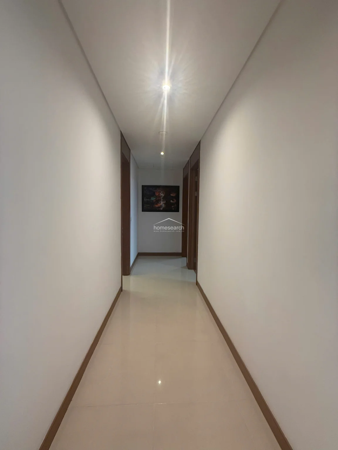This interior hallway features a minimalist, clean design with bright white walls and polished, light-colored tile flooring. The space is illuminated by recessed ceiling lights and leads directly to a framed piece of wall art, creating a simple yet elegant transition between rooms.