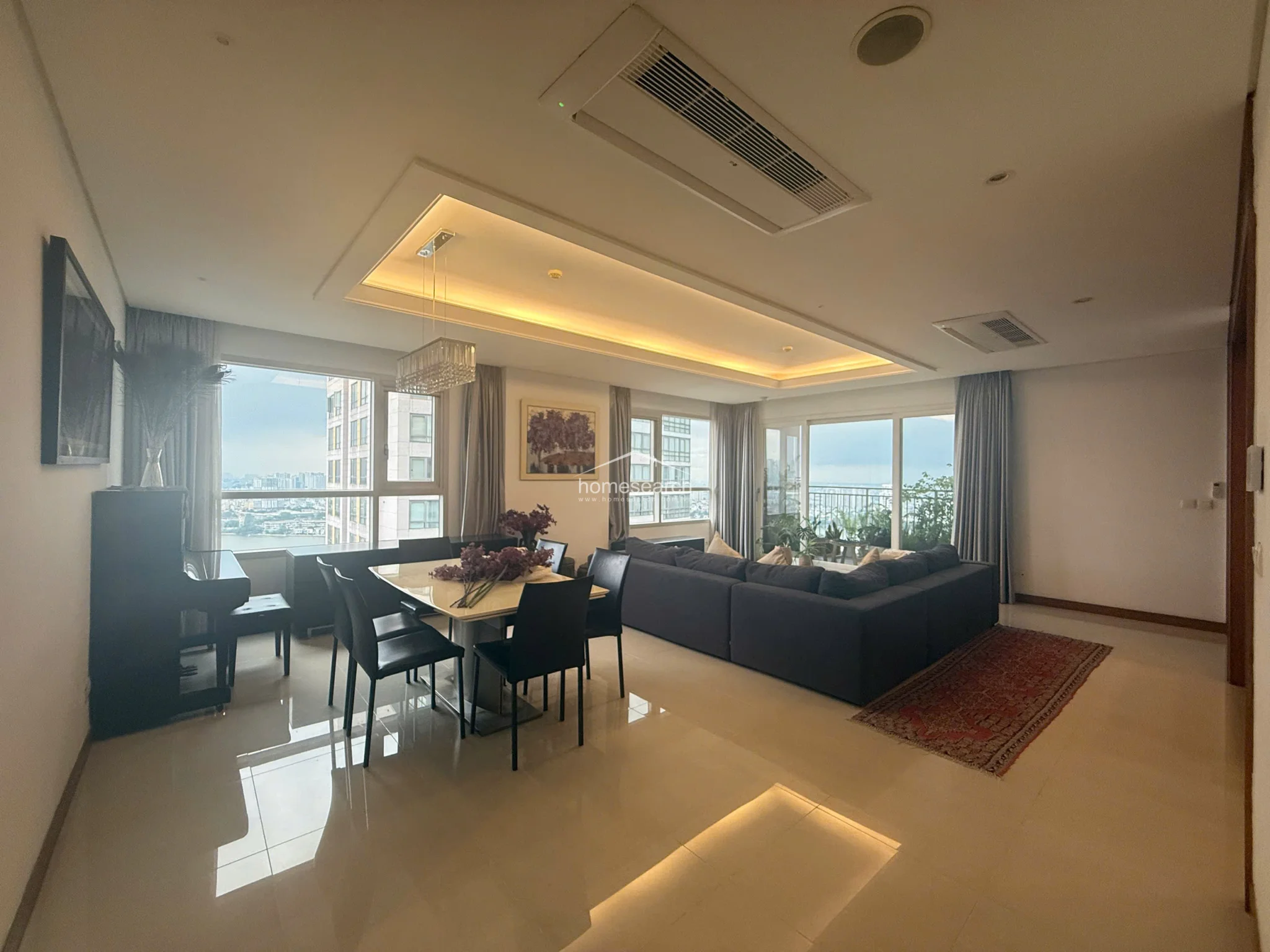 The living room is anchored by a large, dark-toned sectional sofa and a traditional area rug, contrasting against the bright, neutral wall palette. The room is equipped with a modern ceiling-mounted AC unit and large floor-to-ceiling windows that offer an expansive, elevated view of the surrounding skyline and river.
