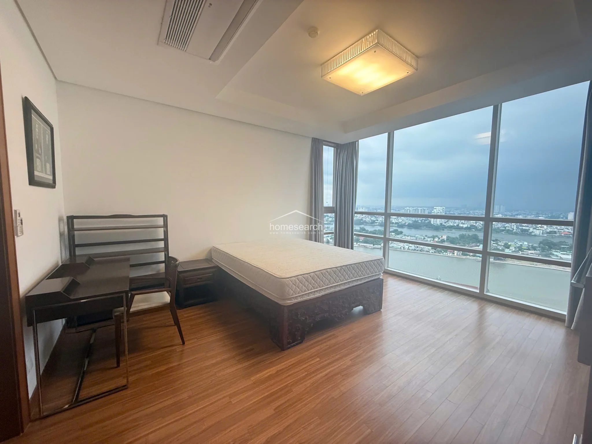 This bedroom features floor-to-ceiling windows that provide expansive, panoramic river and city views, filling the space with abundant natural light. The room is finished with warm wood-look flooring and a neutral color palette, complemented by a ceiling-mounted AC unit and a modern square light fixture. A dark wood bed frame and a matching desk setup provide a classic contrast to the minimalist, clean-lined interior.