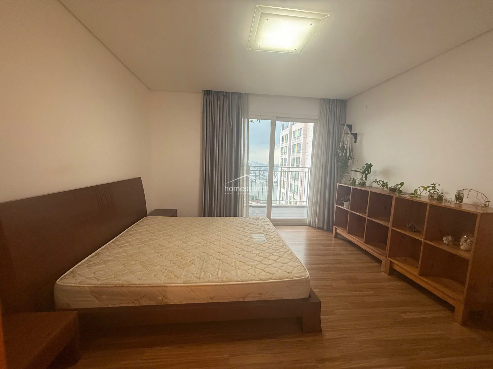 This bedroom offers a clean, minimalist layout centered around a dark wood platform bed, with a large open-shelf storage unit providing ample display space. A sliding glass door leads directly to a private balcony, allowing for excellent natural light and ventilation, while the neutral color palette and wood flooring maintain a consistent, modern aesthetic.
