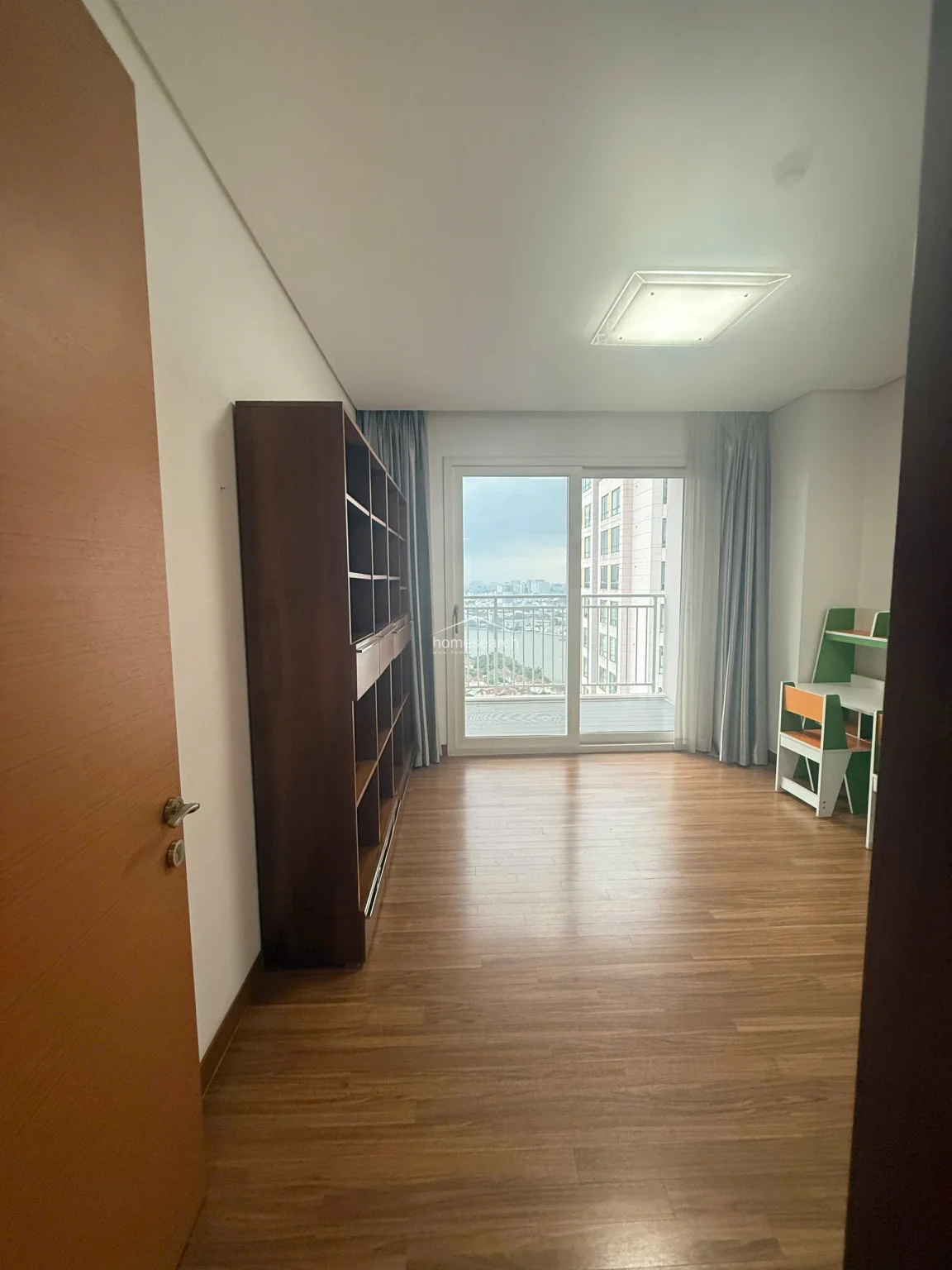 This versatile room is currently configured as a study or secondary bedroom, featuring a tall, dark wood bookshelf and a compact children's desk set. The space connects to a balcony through a sliding glass door, ensuring the room remains bright and functional with a consistent wood-flooring finish throughout.