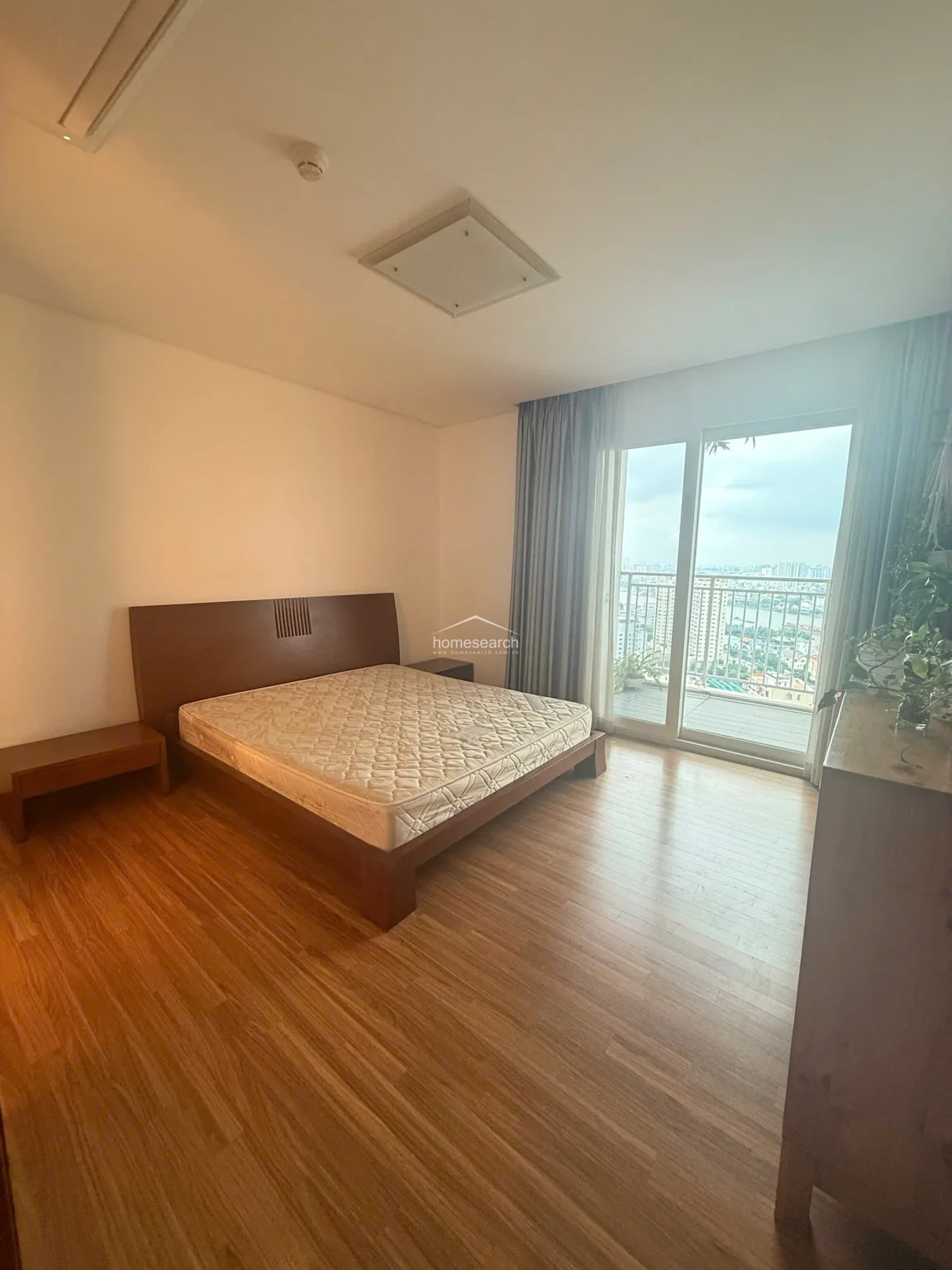 This bedroom maintains the same bright, airy feel as the others, featuring a large sliding glass door that opens onto a balcony with expansive water views. The room is sparsely furnished with a dark wood bed frame and side tables, highlighting the high ceilings and clean, well-maintained wood flooring.