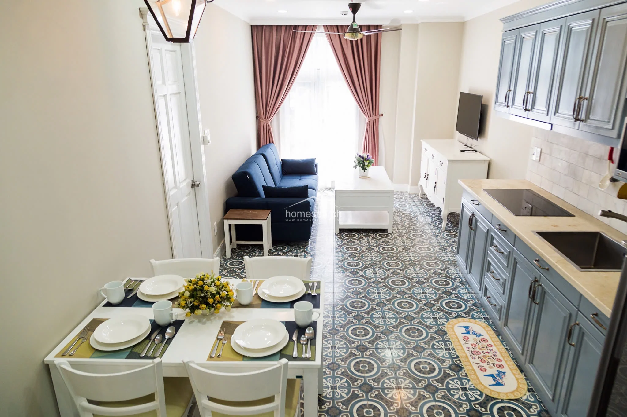 This open-plan living and dining area features a cohesive, light-filled layout with decorative patterned tile flooring that adds a distinct character to the space. The kitchen is equipped with blue-grey cabinetry and a stone countertop, while the living area is anchored by a navy sofa and a large window draped in soft pink curtains, providing ample natural light.