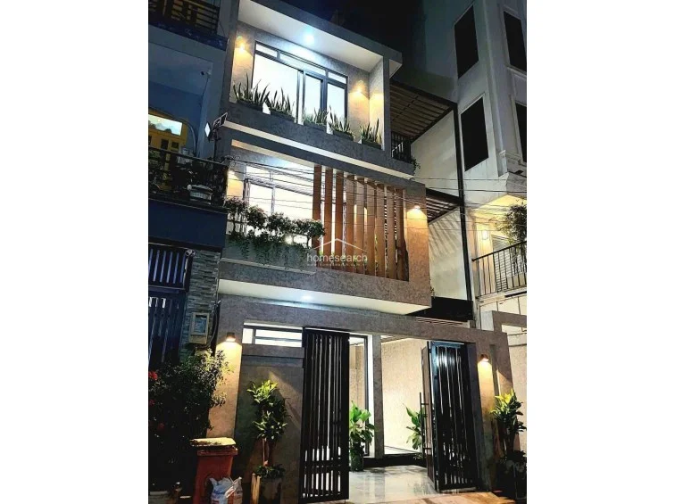 This evening view highlights the architectural lighting of the townhouse, which accentuates the textured facade and the warm glow from the interior. The combination of vertical wooden slats, lush greenery in the balcony planters, and strategic exterior sconces creates a welcoming and modern curb appeal.