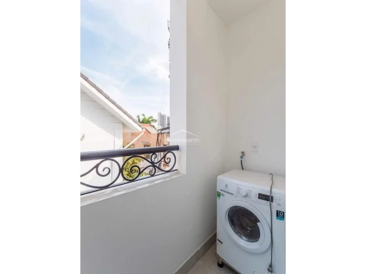 This compact balcony serves as a dedicated laundry space, equipped with a front-loading washing machine and plumbing connections. It features a decorative wrought-iron railing and offers an open-air view of the surrounding neighborhood buildings.