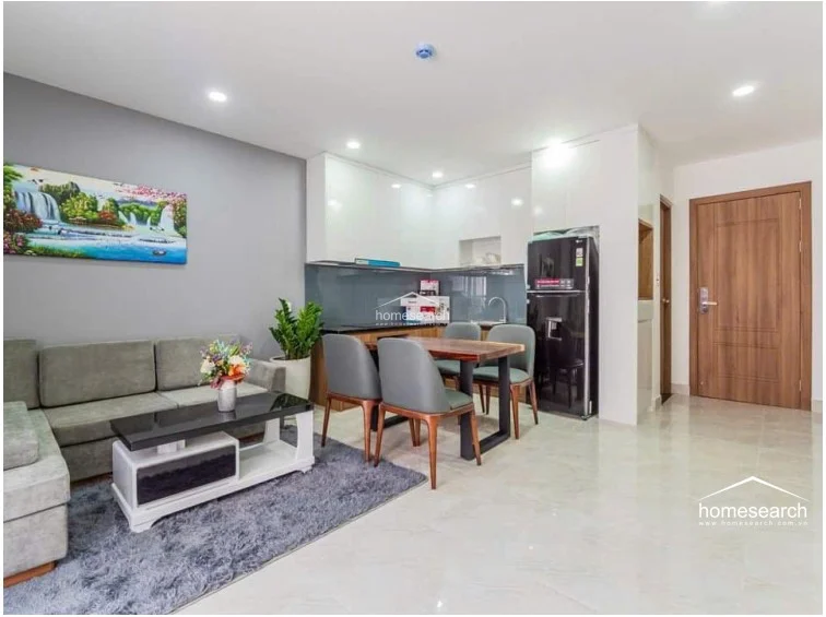 The living area is furnished with a grey sectional sofa and a sleek coffee table, flowing directly into the dining space and kitchen. The room is maintained in excellent condition, featuring a contemporary aesthetic with minimalist decor and an integrated air conditioning unit.