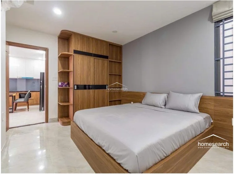 This bedroom features a modern, minimalist design with a warm wood-toned built-in wardrobe and matching bed frame. The room is finished with polished light-colored tile flooring and a neutral gray accent wall, while a doorway provides a direct, open flow into the adjacent kitchen area.
