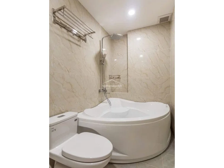 This bathroom features a unique, space-saving corner soaking tub equipped with a handheld shower fixture. The room maintains a consistent aesthetic with light marble-patterned wall tiles and includes a wall-mounted towel rack and a ceiling-integrated exhaust vent.
